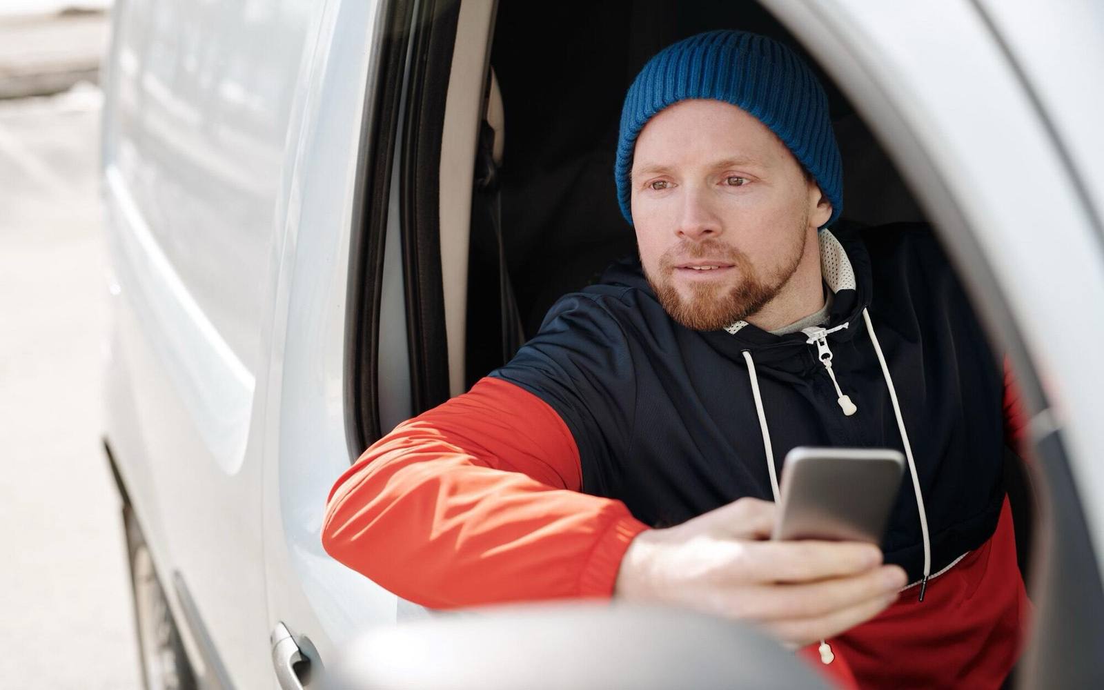 Ein Fahrer mit blauer Mütze lehnt aus dem Fenster eines weißen Transporters und hält sein Telefon in der Hand.