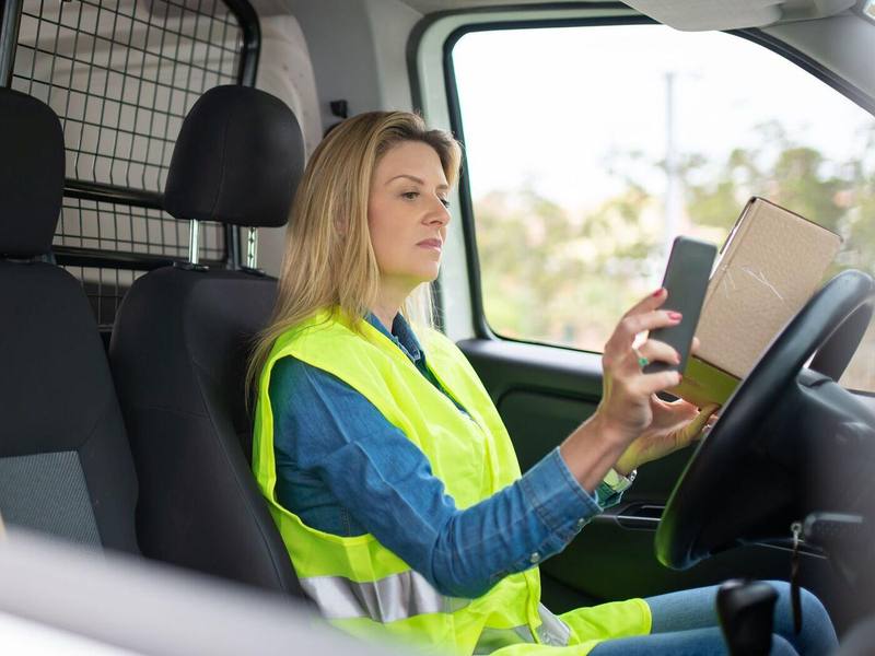 Eine Fahrerin mit Warnweste sitzt am Steuer eines Transporters und schaut auf ihr Telefon.