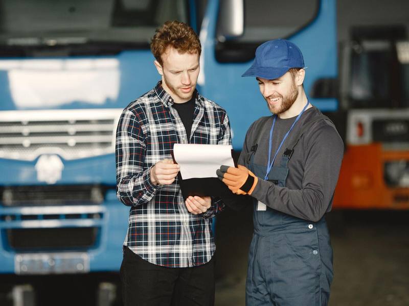 Ein Fahrer im karierten Hemd und ein Kollege in Arbeitskleidung sehen sich vor einem blauen Lkw gemeinsam ein Klemmbrett an.
